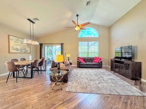 A spacious living room with a dining area, a ceiling fan, a large window, and a television.