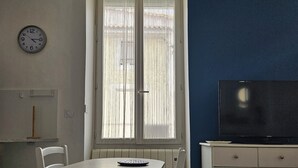 Huoneessa on ruokapöytä, tuoleja, kello, televisiota sisältävä kaappi ja ikkuna, jossa on verhot.