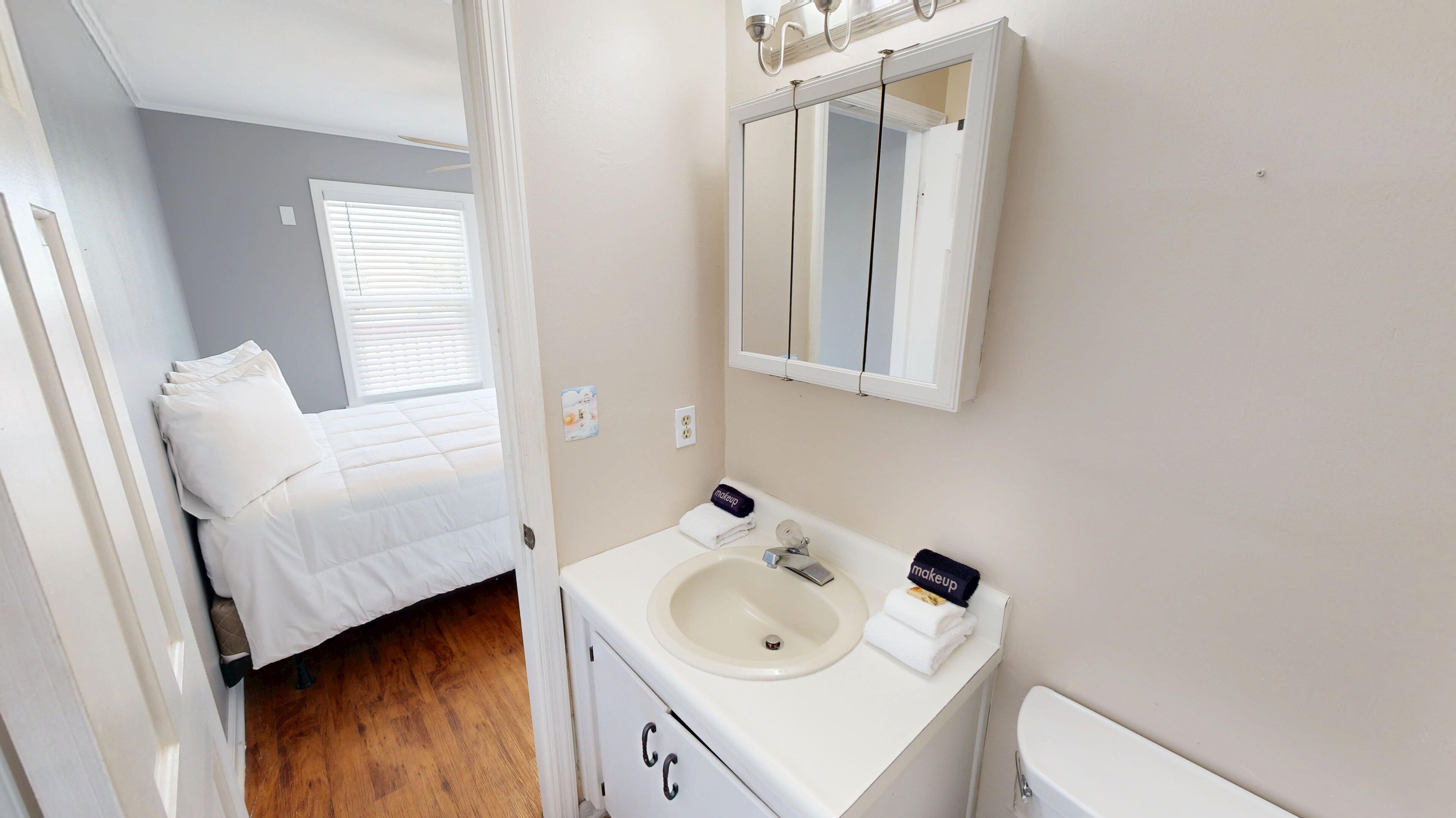 Un dormitorio con cama, un baño con lavamanos y espejo, y una ventana con persianas.