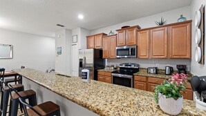 A modern kitchen with granite countertops, wooden cabinets, stainless steel appliances, and bar stools. - Sunny Paradise Retreat At Paradise Palms By Shine Villas 4 Bedroom Townhouse (Kissimmee)