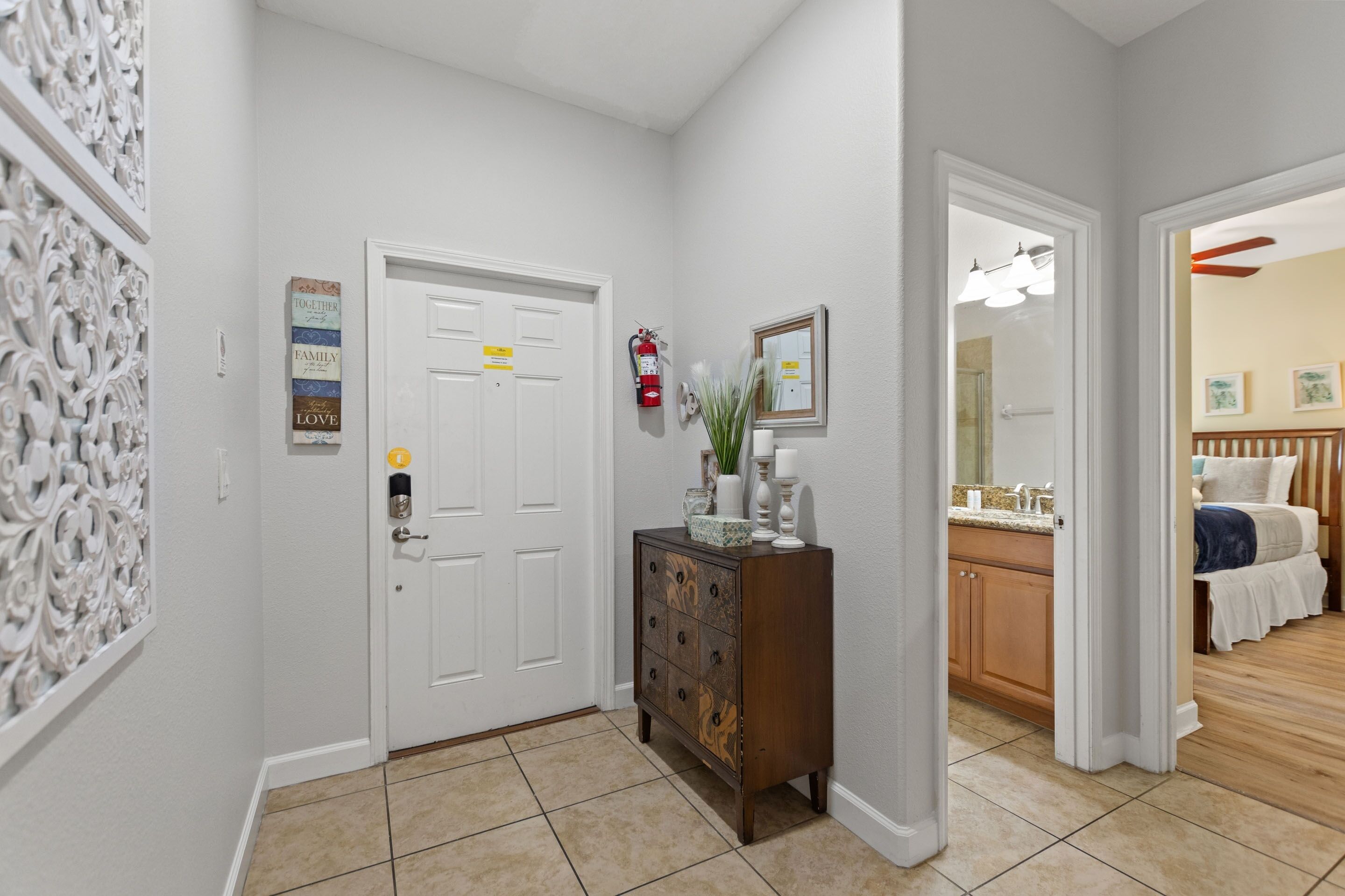 Un couloir avec une porte blanche, un meuble en bois orné d’objets décoratifs et une salle de bain visible à travers une porte entrouverte.