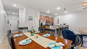 A dining area with a wooden table set for four, a kitchen in the background, and a living area with a sofa and chairs. - Sunny Paradise Retreat At Paradise Palms By Shine Villas 4 Bedroom Townhouse (Kissimmee)