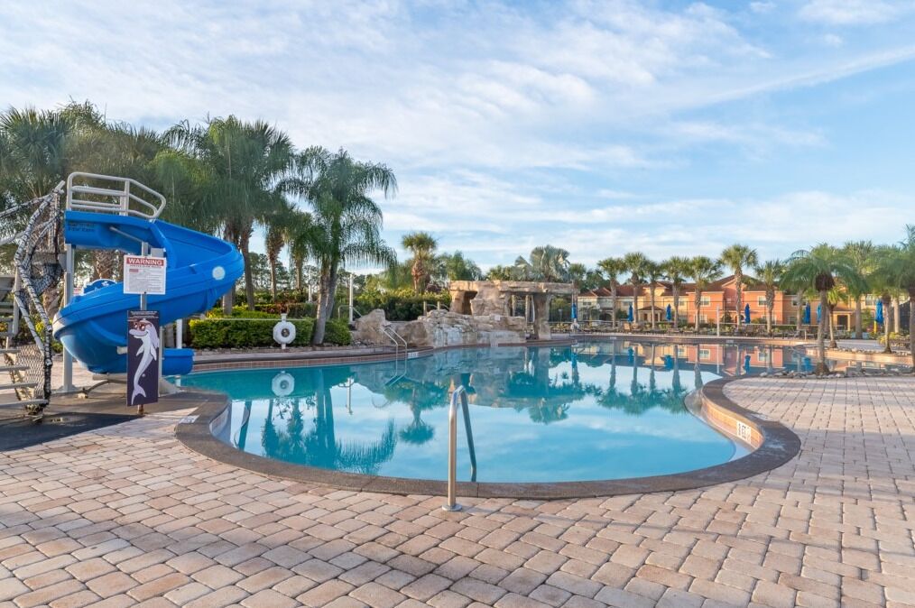 A swimming pool with a blue water slide, palm trees, and a building in the background.