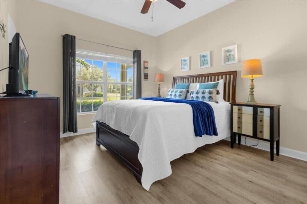 A bedroom with a bed, a nightstand, a lamp, a window with curtains, and a ceiling fan.