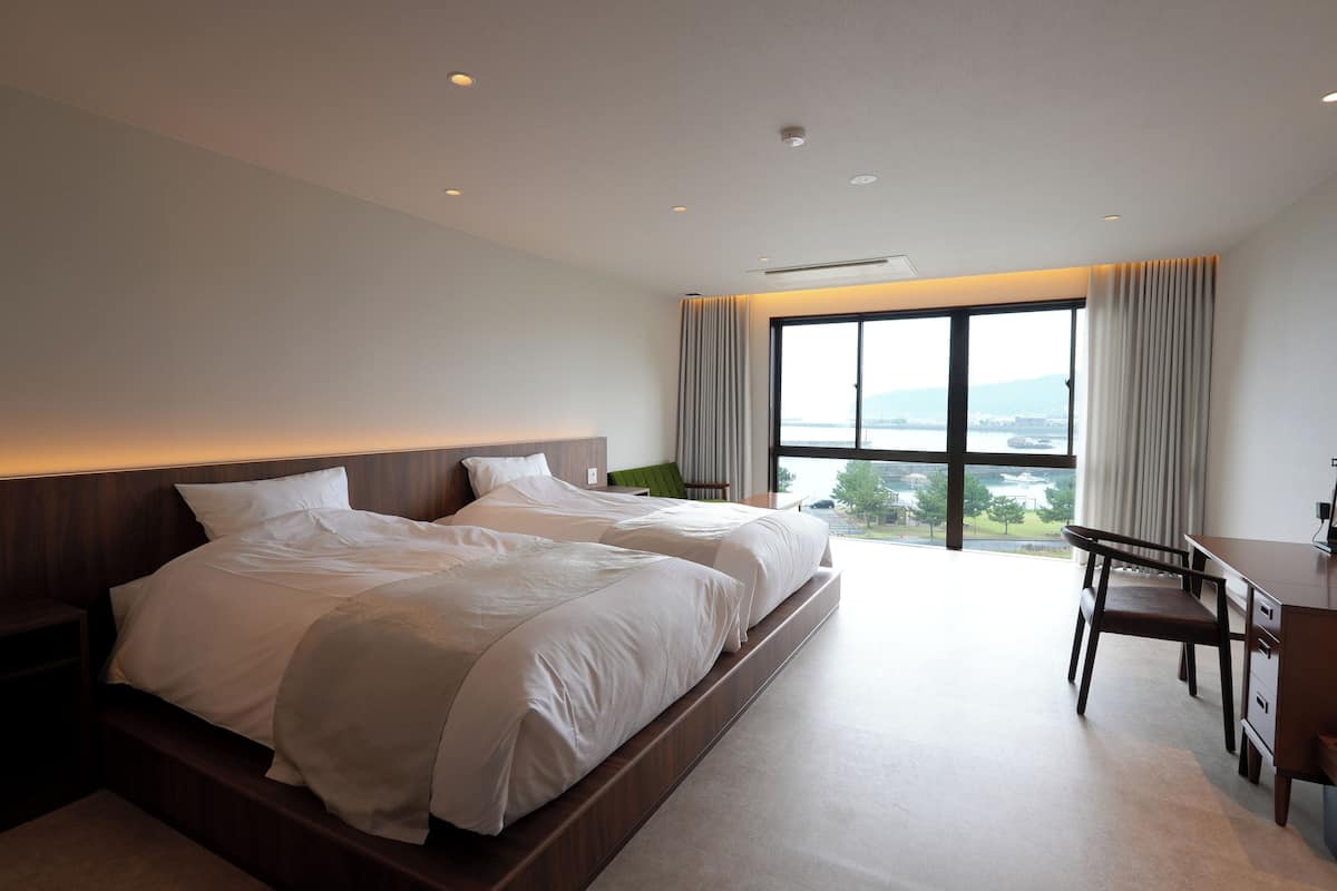 A modern hotel room with two beds, a desk, and a view of the city.