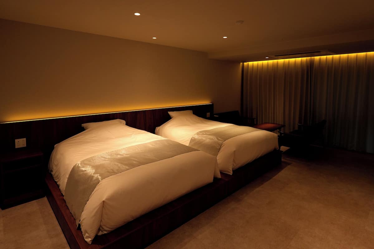 A hotel room with two beds, a nightstand, a lamp, and a curtain.