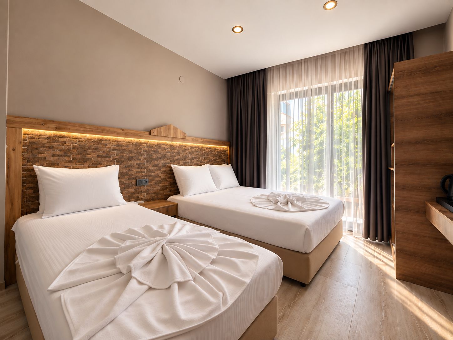 Habitación de hotel con dos camas, cabecera de ladrillo, ventana grande con cortinas y armario de madera.