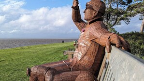 A bronze statue of a seated figure with raised arms, overlooking a grassy area with a view of the sea.
