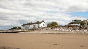 A white building with multiple chimneys, situated on a rocky shoreline with a sandy beach in the foreground.