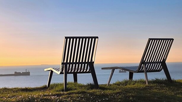 Two metal chairs with slatted backs facing a body of water and a distant bridge.