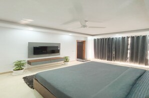 A modern bedroom with a large bed, a flat-screen TV mounted on the wall, a ceiling fan, and a window with curtains. - Housr Sector 56 Gurgaon Golf Course Road (Gurugram)