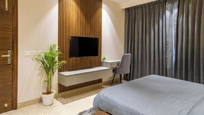Quarto de hotel moderno com uma cama grande, televisão presa a uma parede de painel de madeira, escrivaninha com cadeira e um vaso de planta.