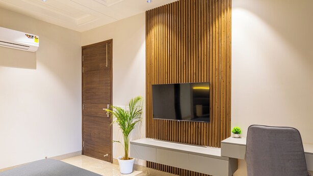 Quarto moderno com uma cama grande, uma TV fixada em uma parede de painéis de madeira, uma escrivaninha com cadeira e um vaso de planta.