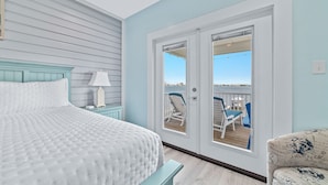 A bedroom with a bed, a nightstand, a lamp, and a view of an outdoor deck with chairs.