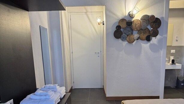 A modern hotel room with a bed, a bedside table with towels, a wall-mounted light fixture, and a decorative wall art piece. - The better (San Giorgio a Cremano)