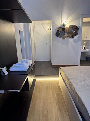 A modern hotel room with a bed, a bedside table with towels, a wall-mounted light fixture, and a decorative wall art piece. - The better (San Giorgio a Cremano)