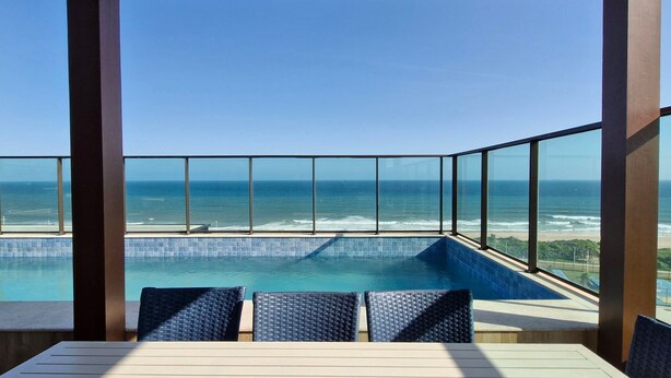 Un espace aménagé au bord de la piscine, offrant une vue sur l’océan, avec une balustrade en verre et une table en bois entourée de chaises.