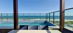 Zona de piscina con vista al océano, con barandilla de cristal y mesa de madera con sillas.