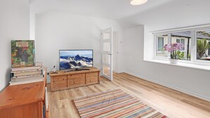 Amplia habitación con piso de madera, un televisor de pantalla plana sobre un mueble de madera, un tapete colorido a rayas y una ventana con vista a un jardín.