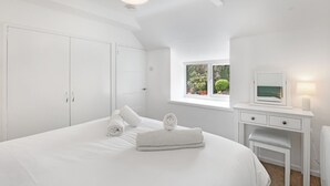Un dormitorio blanco con una cama grande, una mesita de noche con espejo y una ventana con vistas a la vegetación.