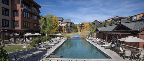 A pool area with lounge chairs and umbrellas, surrounded by buildings with balconies and a clear sky.