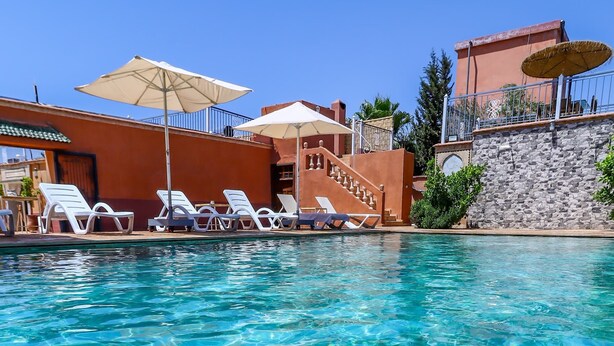 A clear swimming pool with lounge chairs and umbrellas, surrounded by buildings with stone walls and a clear blue sky. - Ferme d'hôte Zomorroda (Saâda)