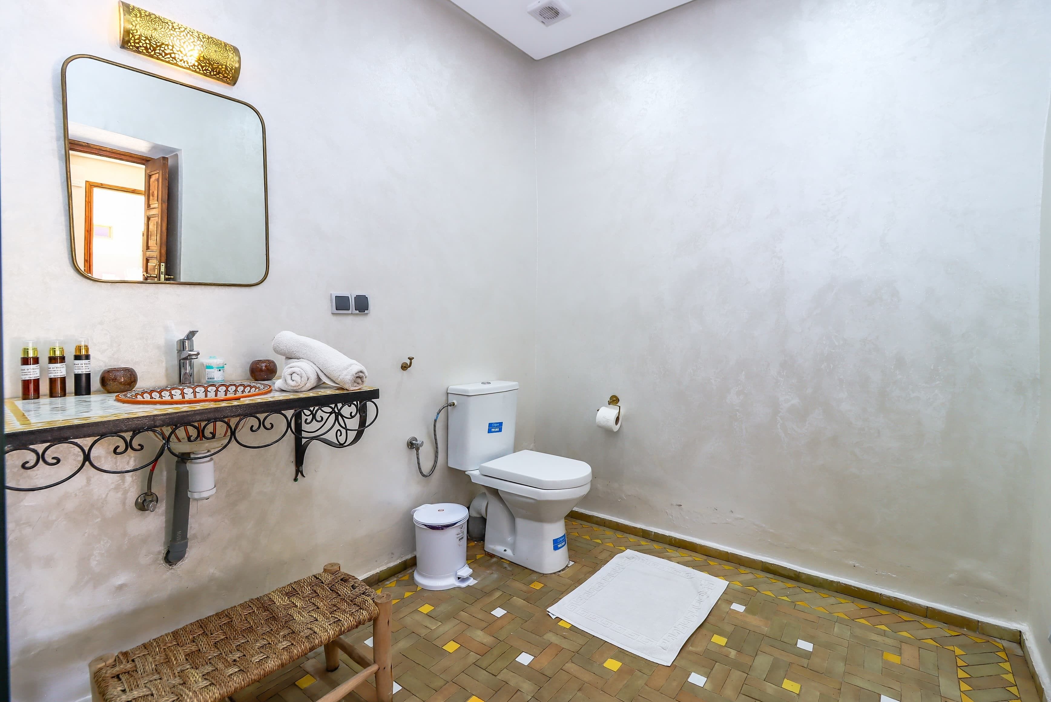 A bathroom with a toilet, sink, and mirror. There is a trash bin and a bench.