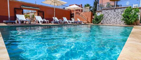 A swimming pool with lounge chairs and umbrellas, surrounded by a building with a stone wall and a staircase.