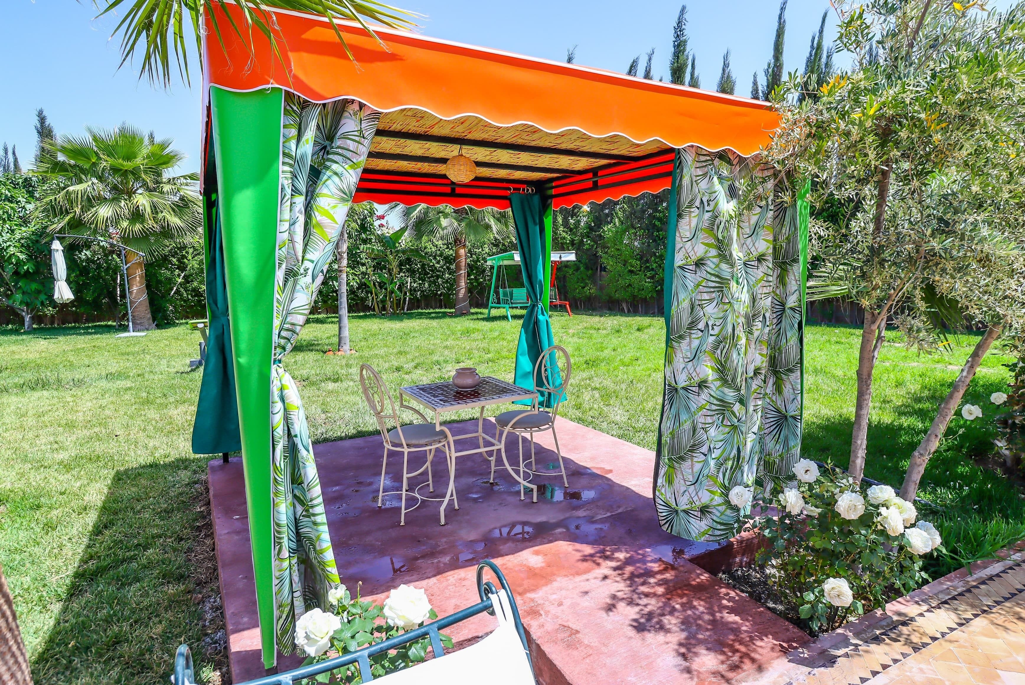 A covered outdoor seating area with a table and chairs, surrounded by greenery and a swing set.