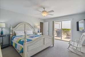 Een slaapkamer met een bed, nachtlampjes, een plafondventilator, een balkon met een stoel en een aan de muur bevestigde televisie.