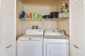 A laundry room with a front-loading washer and dryer, a shelf with cleaning supplies, and a power outlet.