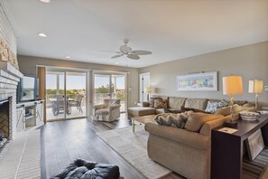 A spacious living area with a sliding glass door leading to an outdoor deck, a comfortable sofa set, a flat-screen TV, and a ceiling fan.