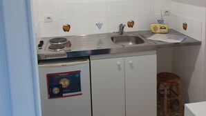 Una pequeña kitchenette con un lavamanos, estufa y radio. Hay un taburete y un armario pequeño.