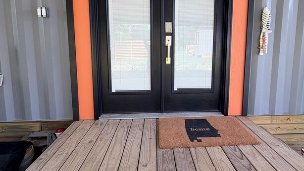Una puerta de marco negro con paneles de vidrio esmerilado, un felpudo con silueta de casa y dos luces fijadas a la pared.