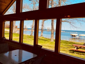 Une cabane dotée de grandes fenêtres donnant sur un lac, avec des tables de pique-nique et une zone herbeuse.