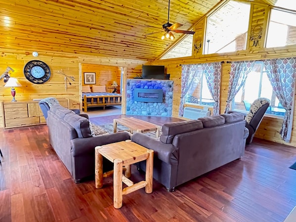 A cozy log cabin interior with a sofa set, a small wooden table, a stone fireplace, and a bed with a blue headboard.