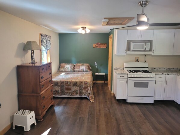 A compact bedroom with a bed, dresser, and a small stool. Adjacent is a kitchenette with a stove, microwave, and cabinets. - Charming one room cabin with direct ATV and Snowmobile trail access (Stewartstown)
