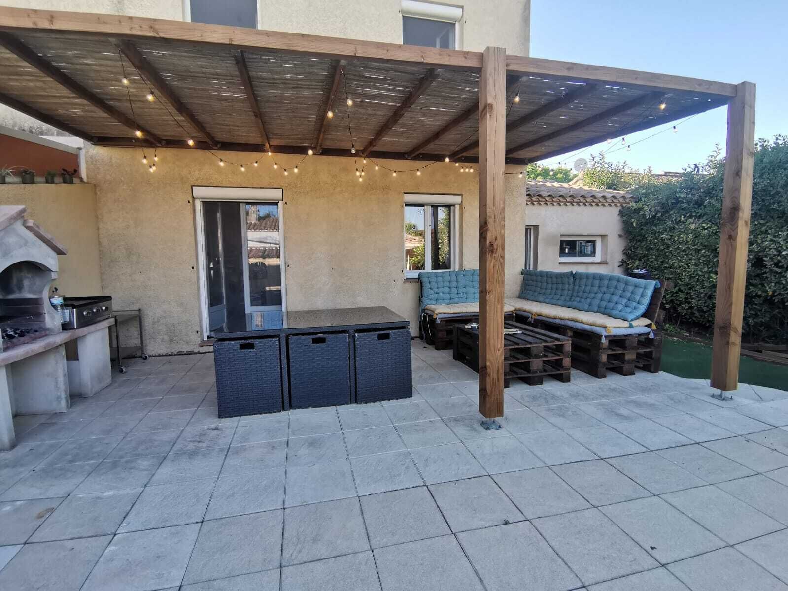 Une terrasse extérieure dotée d’un barbecue, d’une pergola en bois, de guirlandes lumineuses et d’un coin salon bleu avec des coussins.