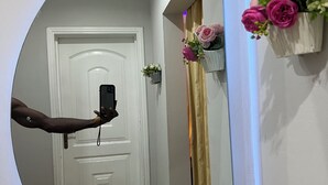 A hallway with a white door, a small potted plant, and a mirror reflecting a person holding a phone. - Luxury 2BR Accra, Near Beach with WiFi, Breakfast, 20min Airport. (Gbawe)
