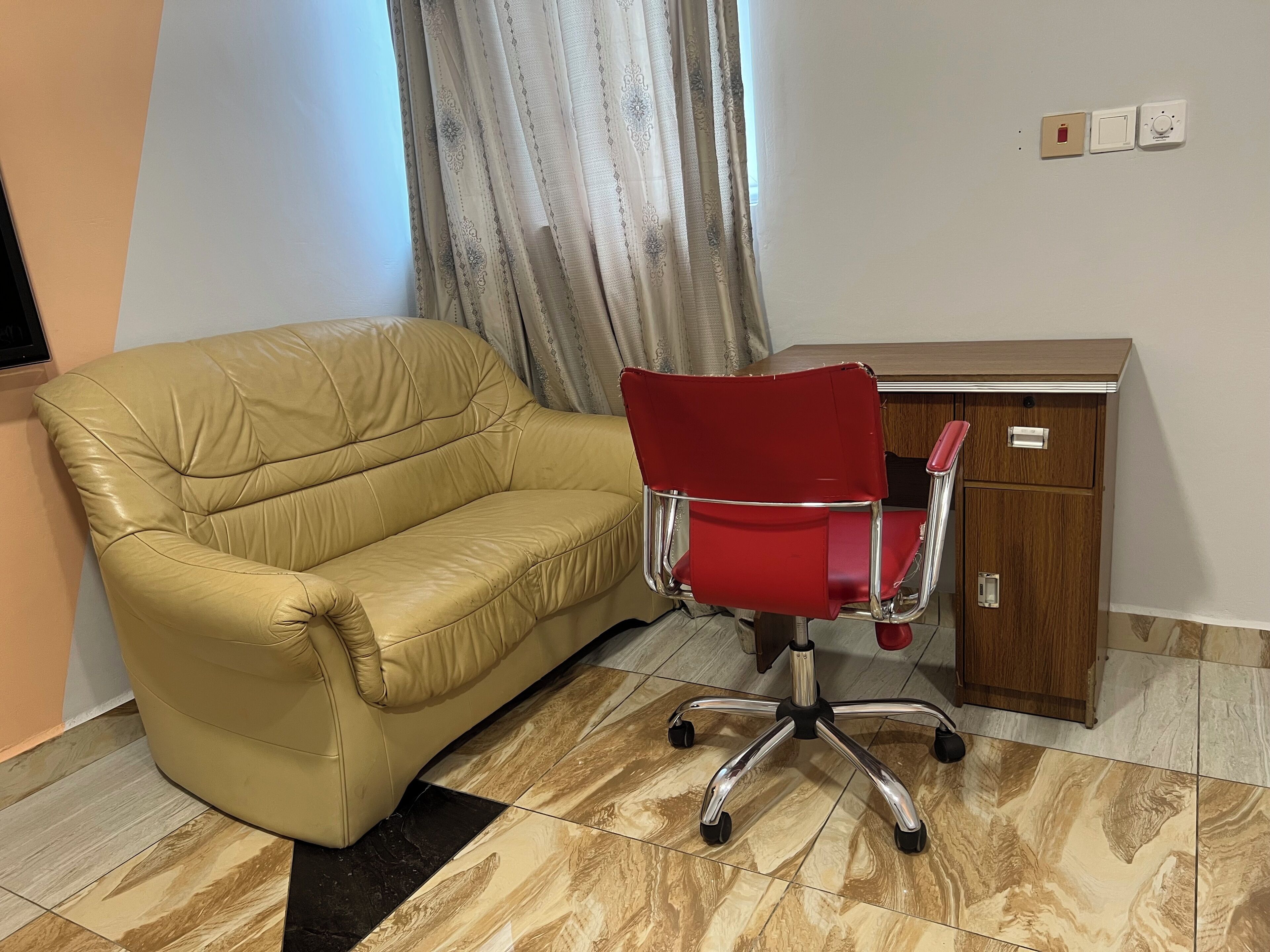 A beige leather sofa and a red office chair in a room with a wooden cabinet and a window with curtains.
