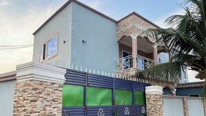 A modern two-story house with a stone facade, a balcony with a metal railing, and a green gate with decorative metalwork. - Luxury 2BR Accra, Near Beach with WiFi, Breakfast, 20min Airport. (Gbawe)