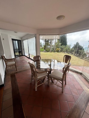 A room with a glass table and chairs, a sliding glass door leading to a garden, and a tiled floor.