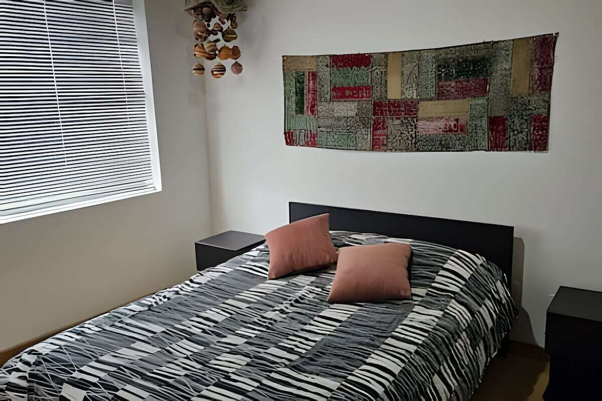 Un dormitorio con una cama, dos mesitas de noche, una ventana con persianas y un cuadro en la pared.