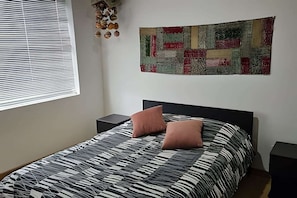 A bedroom with a bed, two nightstands, a window with blinds, and a wall hanging.