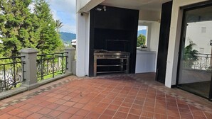 En balkon med grill, flisegulv og udsigt til træer og bjerge.