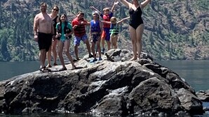 Un grupo de personas sobre un afloramiento rocoso junto a una masa de agua, con montañas al fondo.