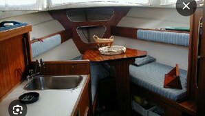 Interior de barco compacto con mesa de madera, lavamanos, una pequeña zona de estar y cajones de almacenamiento.