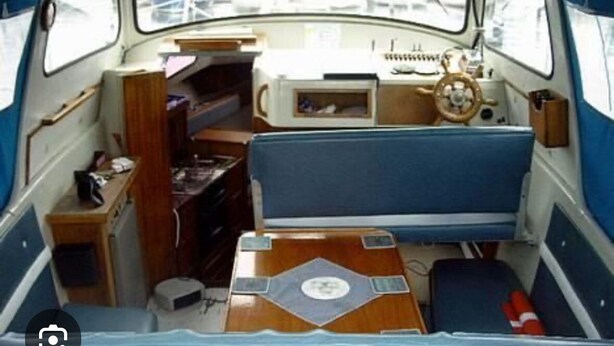 Interior de un barco pequeño con zona de estar central, timón y una mesita.