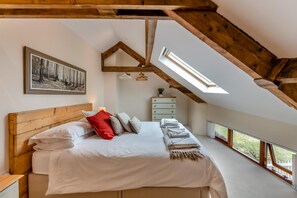 A bedroom with a sloped ceiling, a bed with white bedding, a red pillow, a dresser, and a painting on the wall.