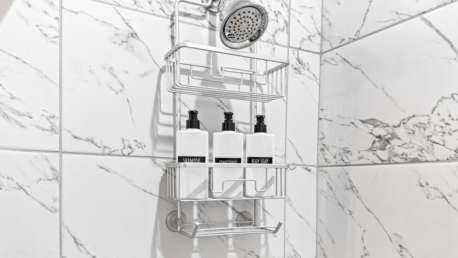 Une salle de bain avec un mur recouvert de carreaux de marbre, une pomme de douche et une étagère en plastique transparent contenant trois bouteilles étiquetées « shampoing », « après-shampoing » et « savon pour le corps ».